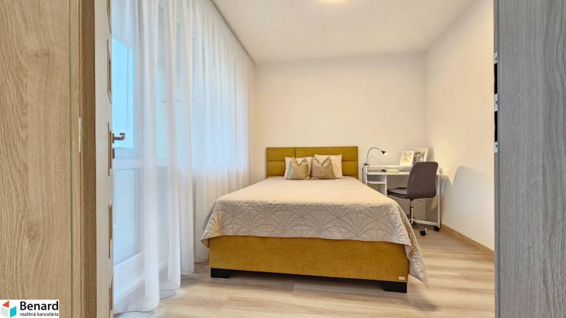 Ložnice v jednopokojovém bytě s postelí, psacím stolem a podlahou s dřevěným dekorem.