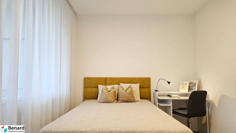 Ložnice v jednopokojovém bytě se světlou postelí, záclonami a bílým stolem s lampou.