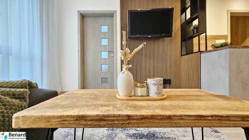 Obývací část 1+kk bytu s dřevěným stolem a dekoracemi na pozadí s televizorem.