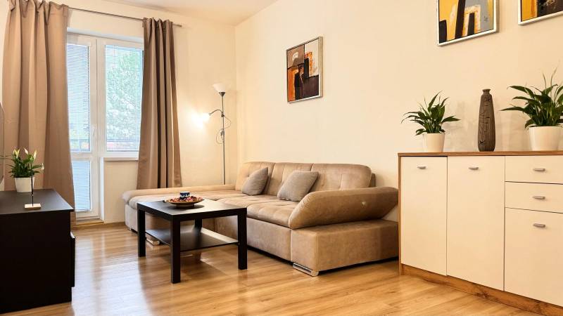 Obývací pokoj v 2pokojovém bytě s béžovou sedačkou, podlaha s dřevěným dekorem.