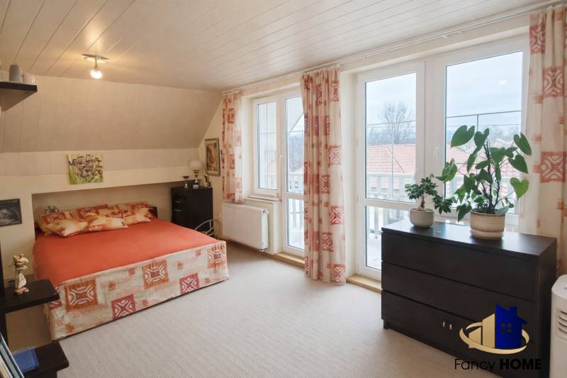 Ložnice v rodinném domě s postelí, skříňkou, velkými okny a rostlinami na parapetu.