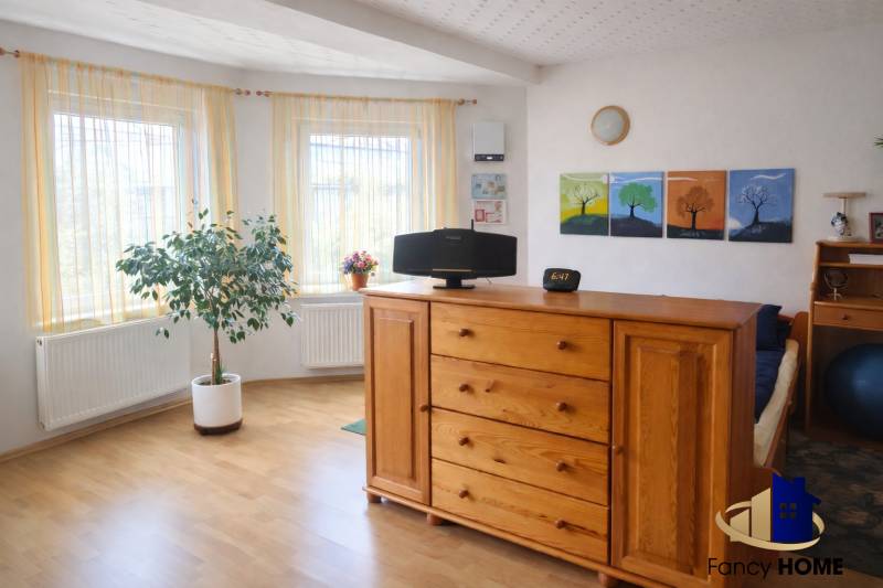 Světlý pokoj v rodinném domě s dřevěným dekorem, komodou a pokojovou rostlinou.