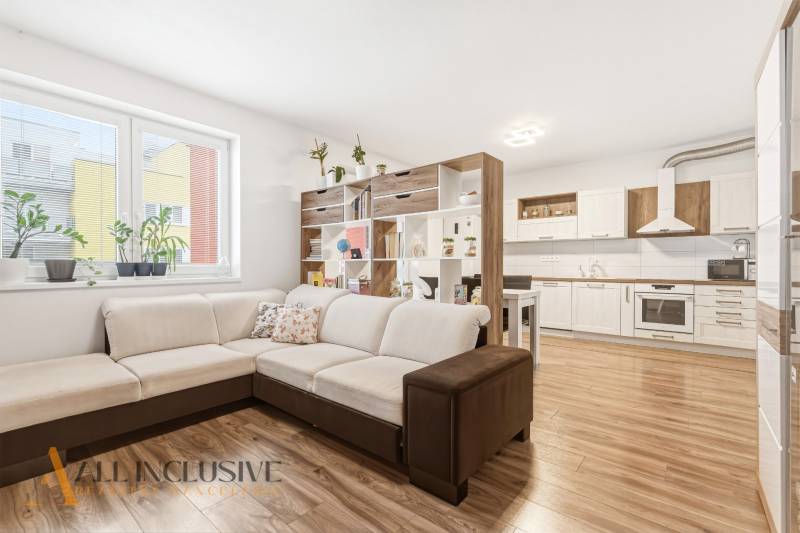 Obývací pokoj v 2-pokojovém bytě s velkou sedačkou a kuchyní, podlaha s dřevěným dekorem.