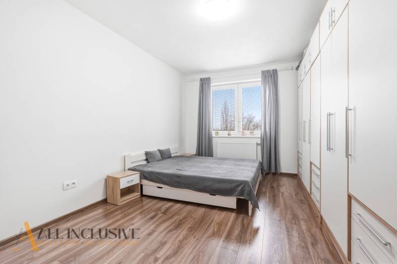 Ložnice v dvoupokojovém bytě s podlahou s dřevěným dekorem, bílými skříněmi a šedými detaily.