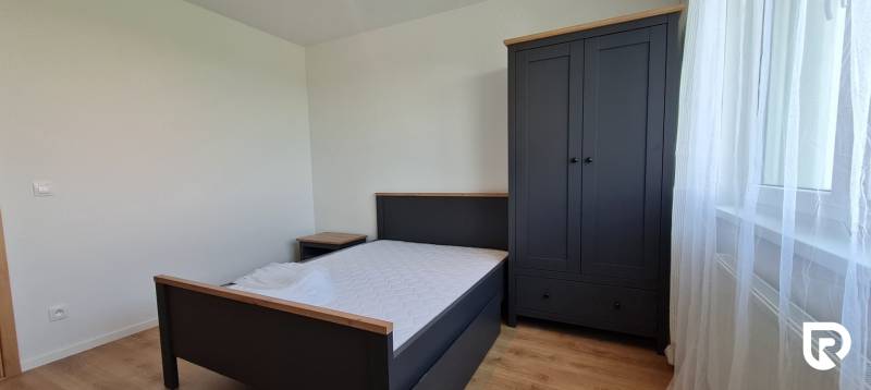 Ložnice v dvoupokojovém bytě obsahuje postel, noční stolek, šatník a podlahu s dřevěným dekorem.