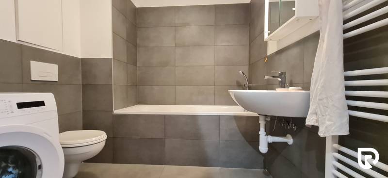 Koupelna s vanou, umyvadlem a pračkou v 2-pokojovém bytě. Šedé obklady dominují prostoru.