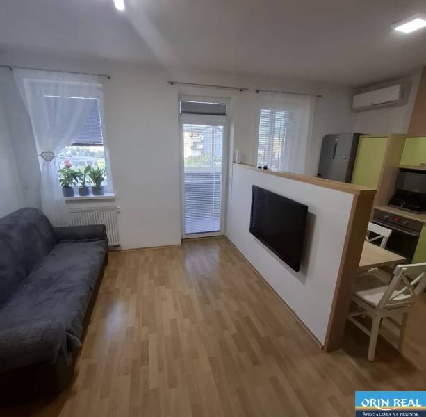 Obývací pokoj dvoupokojového bytu s pohovkou, televizí, podlahou s dřevěným dekorem a balkonem.