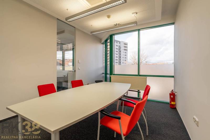 Kanceláře se skleněnou stěnou, bílým stolem a červenými židlemi.