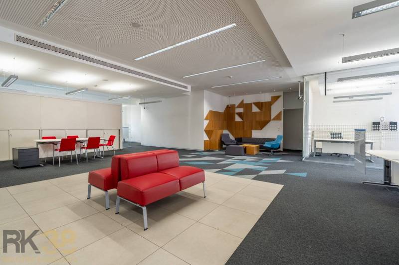 Kanceláře s červenými sedačkami, konferenčním stolem, vzorovaným kobercem a dřevěnými dekoracemi.