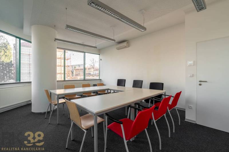 Interiér kanceláře s dlouhým stolem, barevnými židlemi a velkými okny.