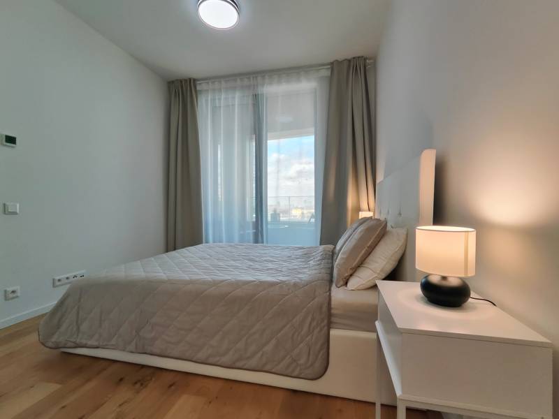 Ložnice v 2pokojovém bytě s postelí, nočním stolkem a podlahou s dřevěným dekorem.