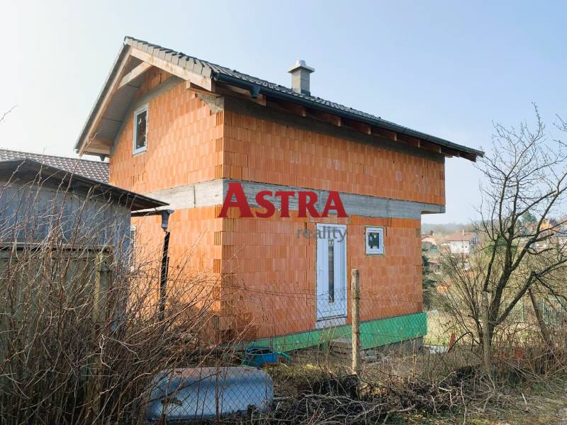 Chata ve výstavbě s cihlovými stěnami u vesnice a zahrady v pozadí.