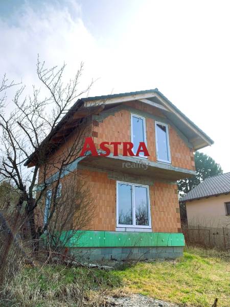 Dvoupatrová chata v rozestavěném stavu, obklopená stromy a trávou na venkovském pozemku.