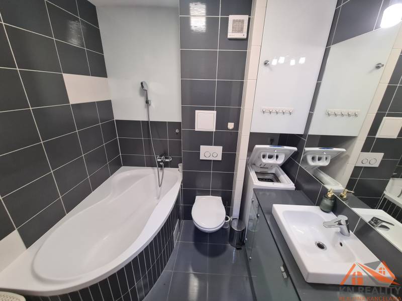 Koupelna v 2-pokojovém bytě s vanou, pračkou, umyvadlem a tmavými obklady.