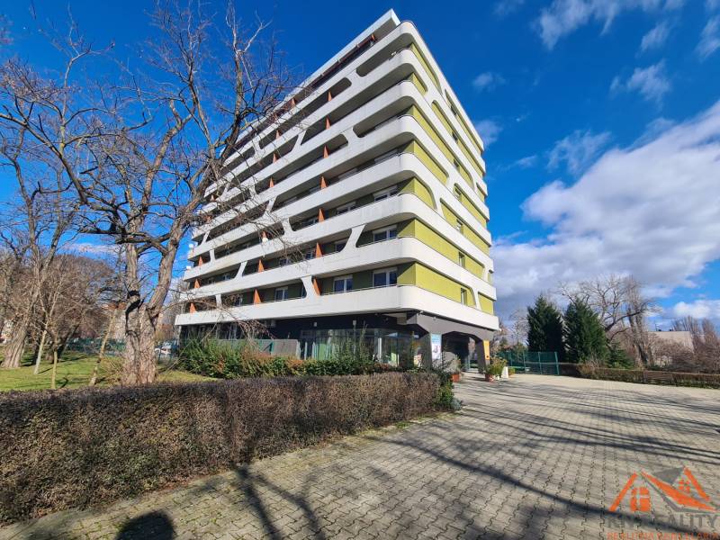 Bytový dům v Komárně se 7 patry, obklopený stromy a dlážděným chodníkem.