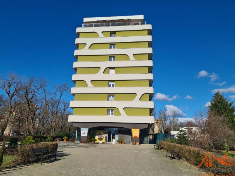 Vícepatrový bytový dům v Komárně s moderním designem, obklopený zelení a lavičkami.