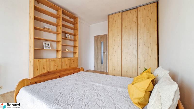 Ložnice v 2pokojovém bytě s postelí, skříní a policemi, podlaha s dřevěným dekorem.