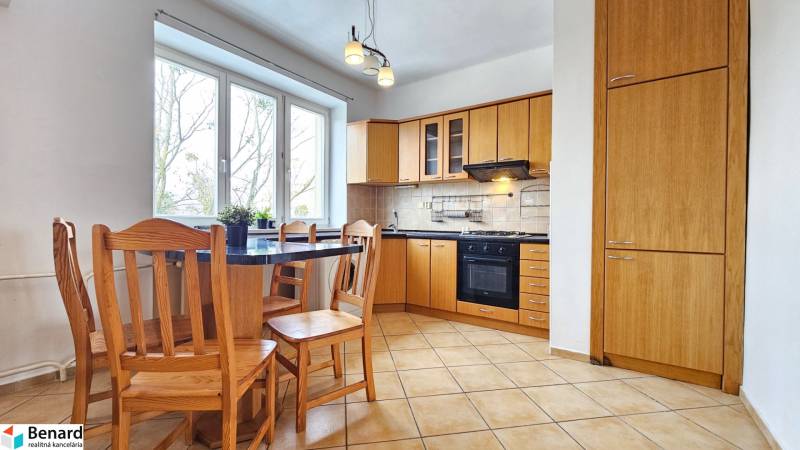 Kuchyně v 2-pokojovém bytě s dřevěným nábytkem a dlažbou, velká okna.