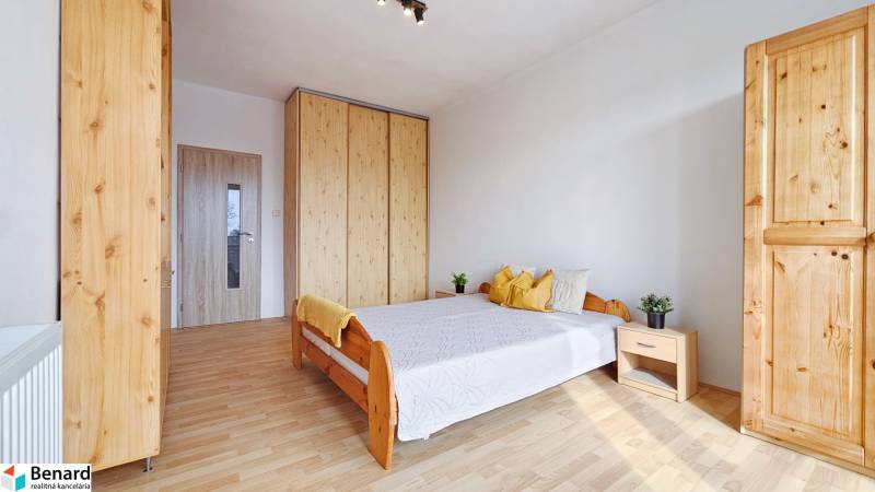 Ložnice v dvoupokojovém bytě s podlahou s dřevěným dekorem, postelí a šatníkovými skříněmi.