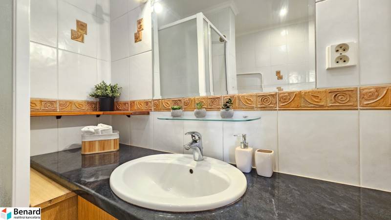 Koupelna v 2-pokojovém bytě s keramickými obklady, umyvadlem a skleněnou policí.