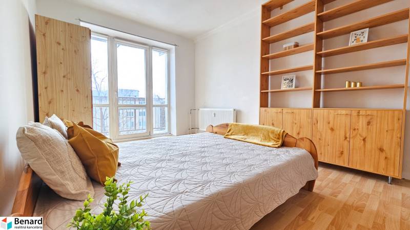 Světlá ložnice v 2-pokojovém bytě s postelí, policovou skříní a podlahou s dřevěným dekorem.