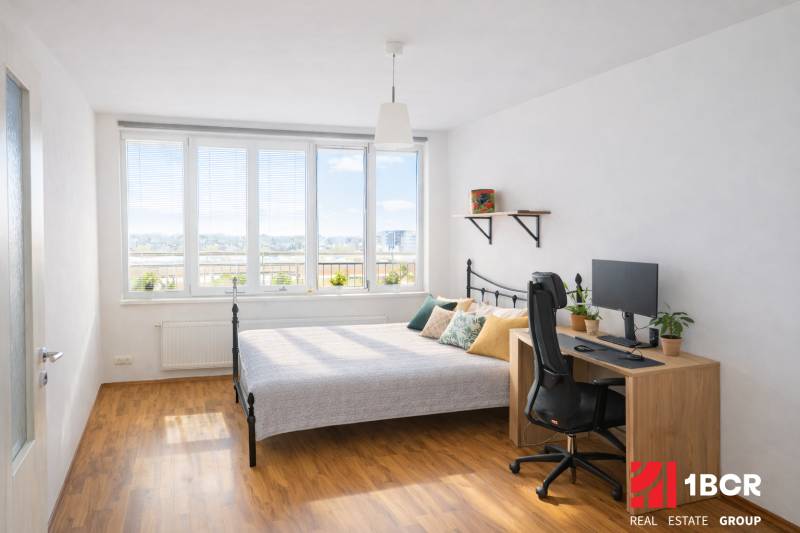 Ložnice v dvoupokojovém bytě s postelí, stolem, počítačem a podlahou s dřevěným dekorem.