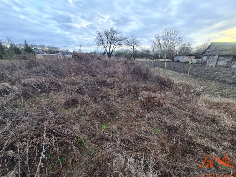 Pozemky - bydlení v Martovcích, neupravený terén, starší dřevěné stavby, stromy, mraky na obloze.