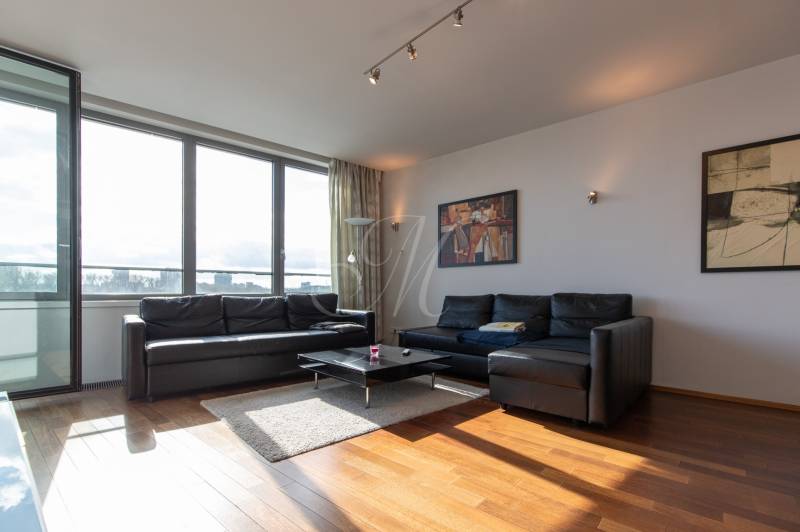 Obývací pokoj v 2-pokojovém bytě s černou sedačkou, podlahou s dřevěným dekorem a velkými okny.