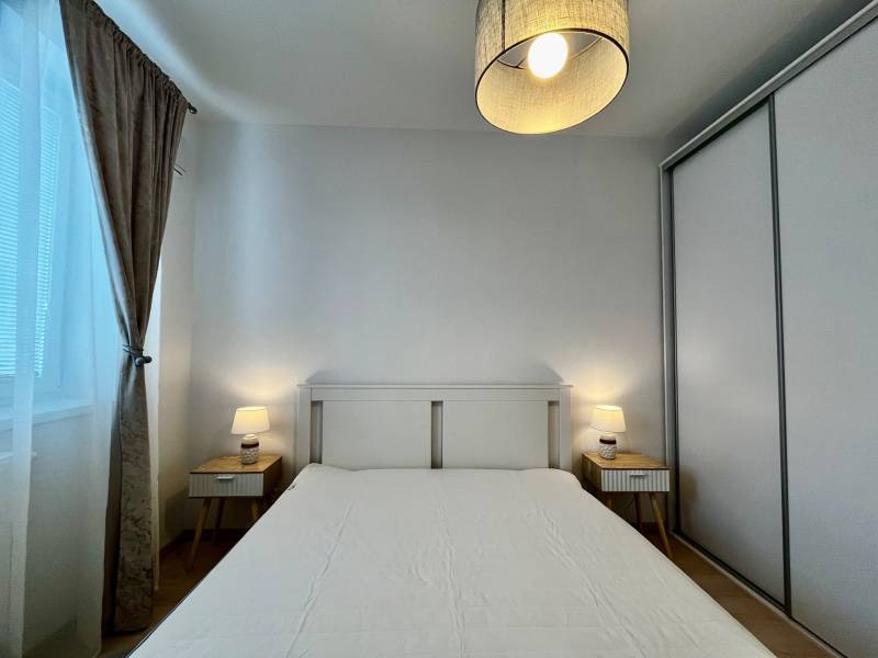 Ložnice v 2pokojovém bytě s bílou postelí, nočními stolky a lampami.