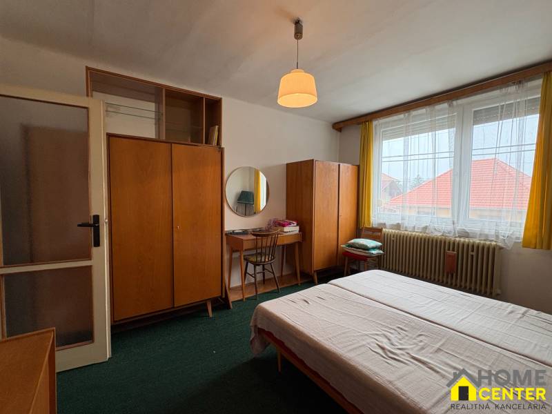 Ložnice rodinného domu s kobercovou podlahou, dřevěným nábytkem a velkým oknem.