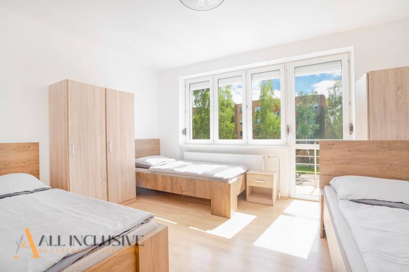 Světlý pokoj se třemi postelemi, bílými stěnami, skříněmi a velkými okny.