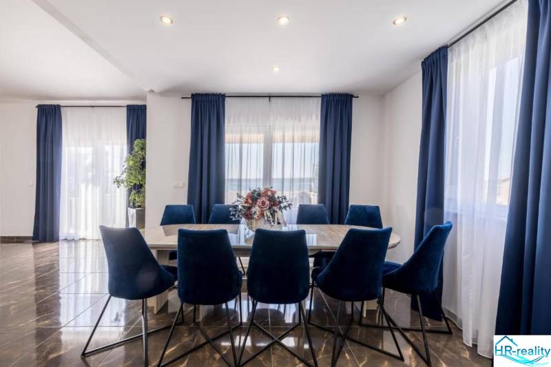 Jídelna ve vile s tmavými závěsy, podlahou s dřevěným dekorem a modrými židlemi.