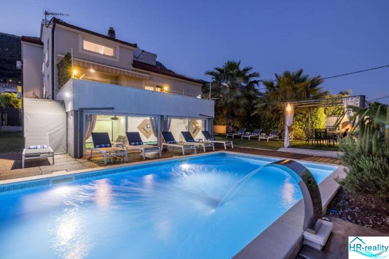 Vila v Kaštela s osvětleným bazénem, lehátky a palmami v zahradě při soumraku.