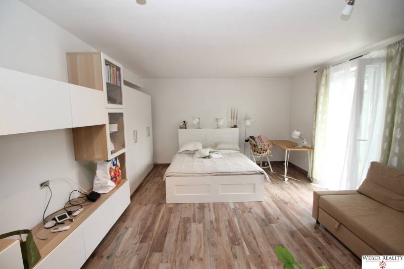 1+kk byt s postelí, sedačkou a skříní, podlaha s dřevěným dekorem.