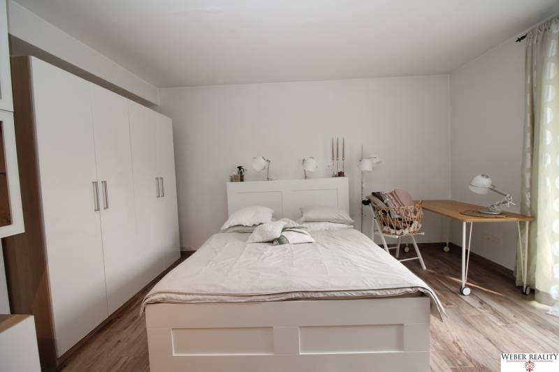 Ložnice v jednopokojovém bytě s postelí, bílou skříní a podlahou s dřevěným dekorem.