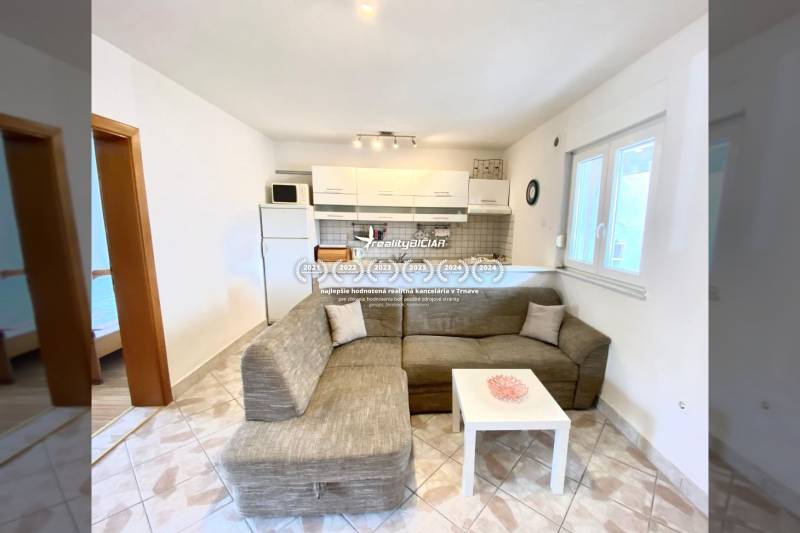 Obývací pokoj v rodinném domě s pohovkou a kuchyňským koutem, dlažba na podlaze.