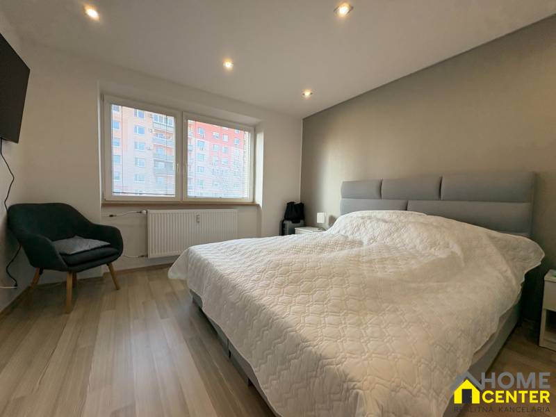 Ložnice v dvoupokojovém bytě s podlahou s dřevěným dekorem, pohodlným křeslem a velkým oknem.