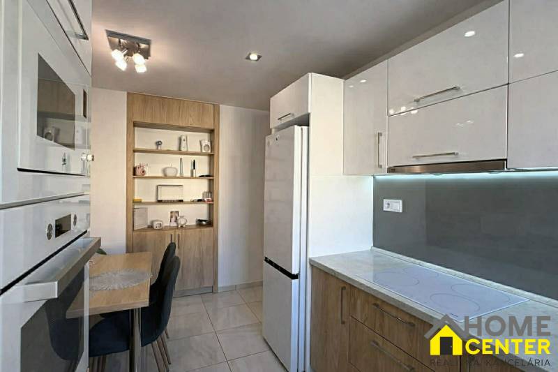 Kuchyně v 2-pokojovém bytě s bílými skříňkami, stolem a dřevěným dekorem polic.