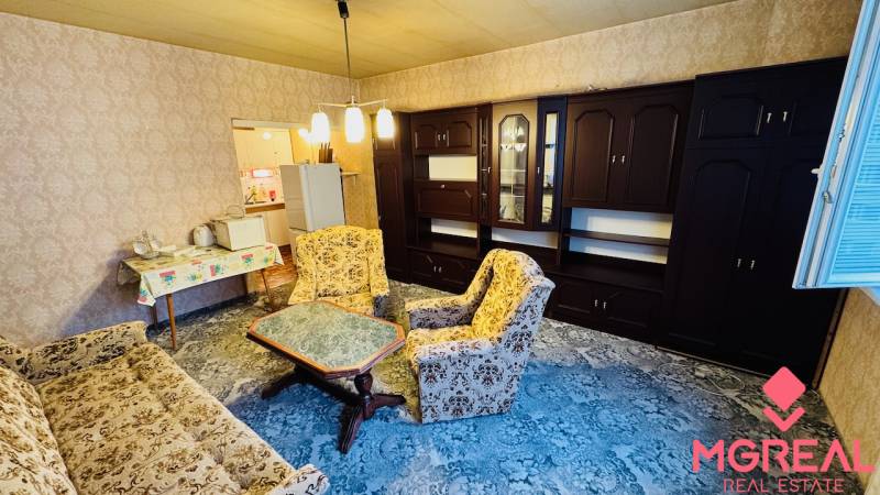 Obývací pokoj v dvoupokojovém bytě se starším nábytkem a kobercem na podlaze.