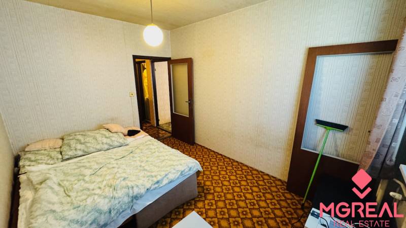 Ložnice s kobercem, postelí a světlými stěnami v dvoupokojovém bytě.