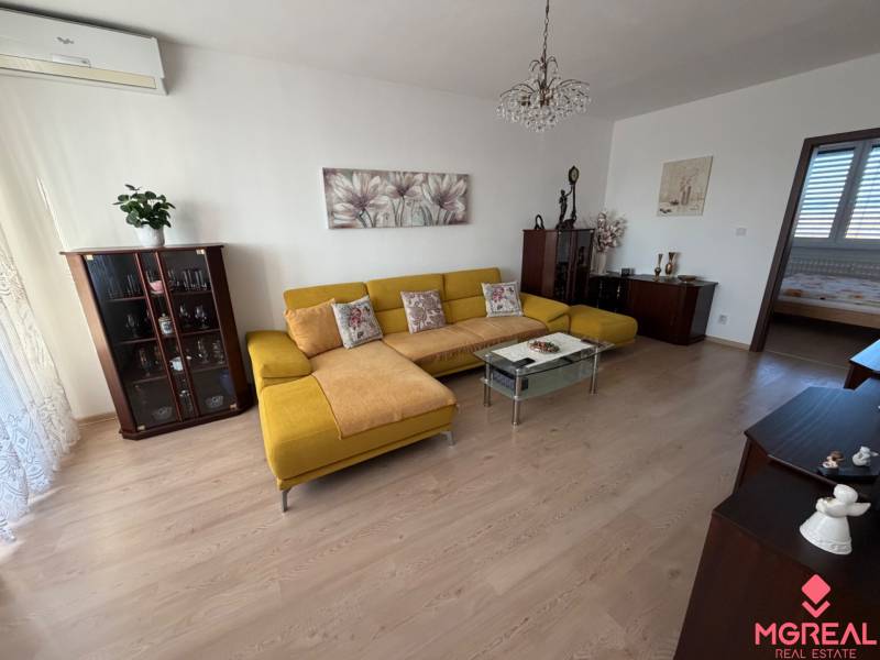 Obývací pokoj s podlahou s dřevěným dekorem, žlutou sedačkou a skleněným stolkem v 3-pokojovém bytě.