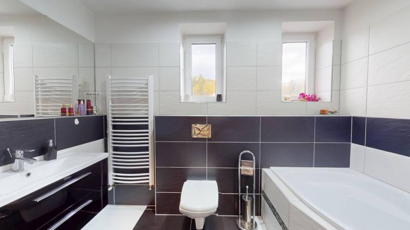 Koupelna v rodinném domě s vanou, dlaždicemi, umyvadlem a okny na stěně.