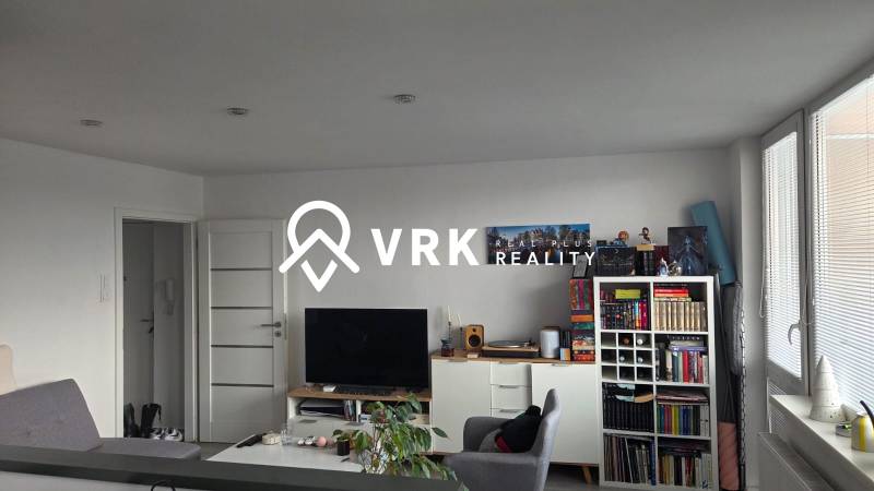 Obývací pokoj třípokojového bytu s knihovničkou, televizorem a světlým nábytkem.