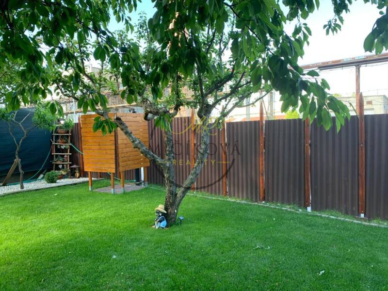 Zahrady v Dolních Krškanech, Nitra, s trávníkem, stromem, malým dřevěným domečkem a plotem.