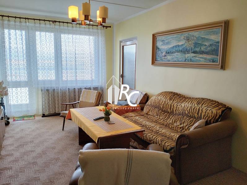 Obývací pokoj s pohovkou, křeslem a stolem v dvoupokojovém bytě s velkým oknem.