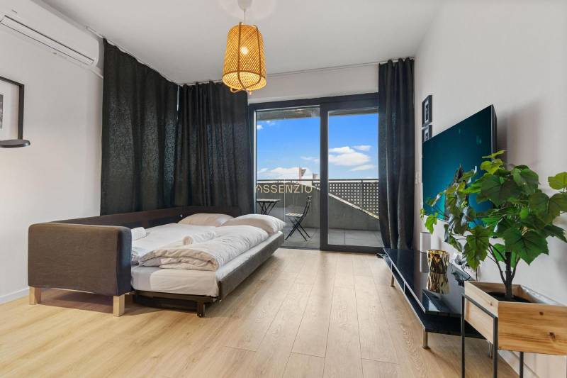 Obývací část v 1-pokojovém bytě s postelí, podlahou s dřevěným dekorem a balkonem.