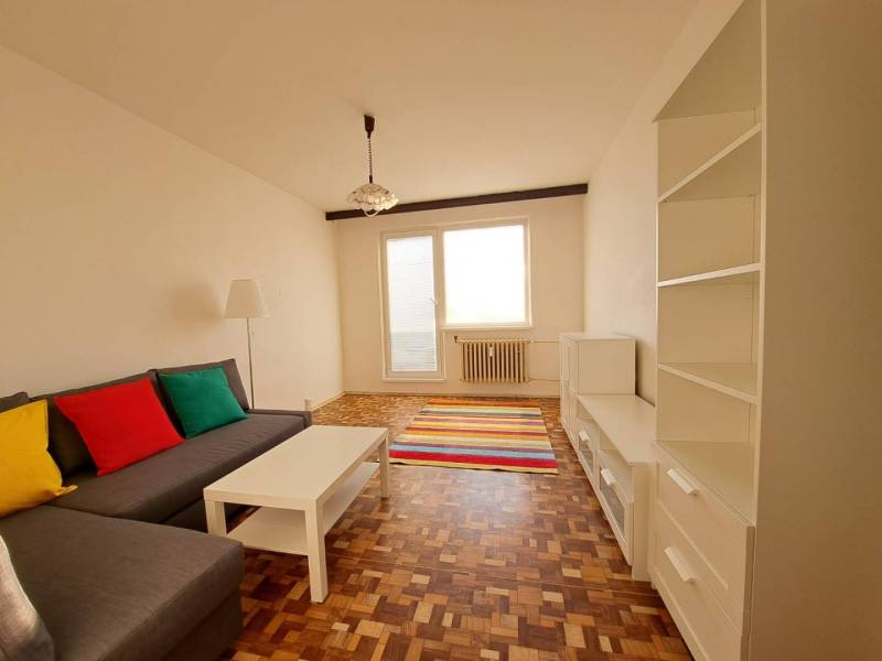 Obývací pokoj v 2-pokojovém bytě s pohovkou, bílým nábytkem, pestrobarevnými polštáři a podlahou s dřevěným dekorem.