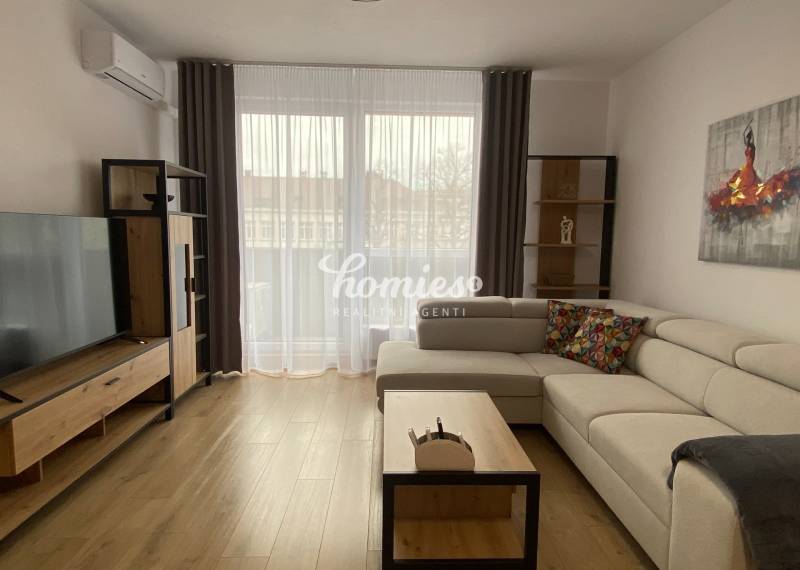 Obývací pokoj v 2pokojovém bytě s podlahou s dřevěným dekorem, světlou sedačkou a televizí.