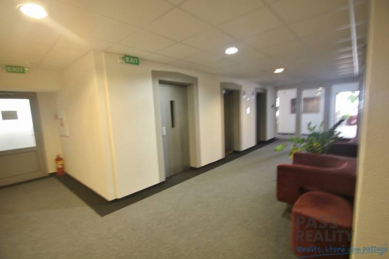 Interiér kanceláře s výtahy a sedačkou, bílá stěna, piktogramy “EXIT”.