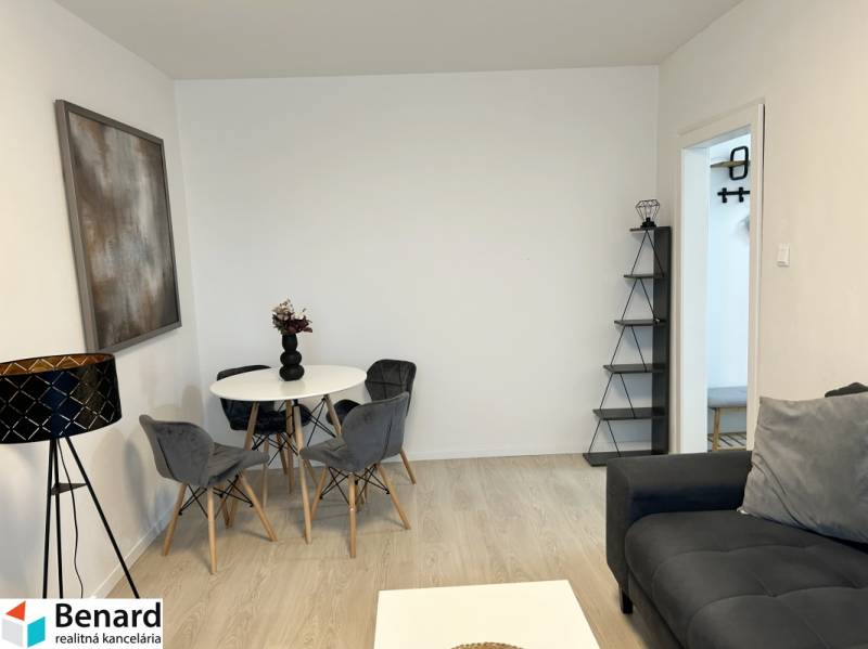 Obývací pokoj v 2-pokojovém bytě s podlahou s dřevěným dekorem, šedou sedačkou a jídelním stolem.